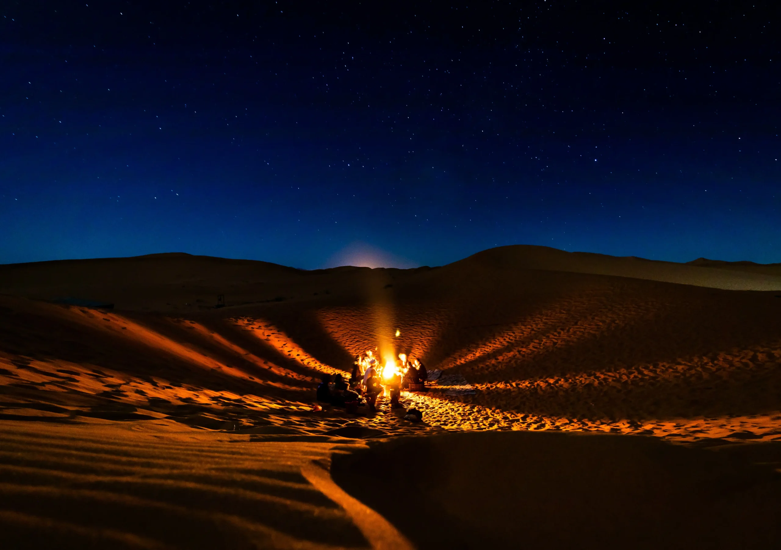 Night in the Sahara Desert with a glowing campfire among sand dunes beneath a star-filled sky.